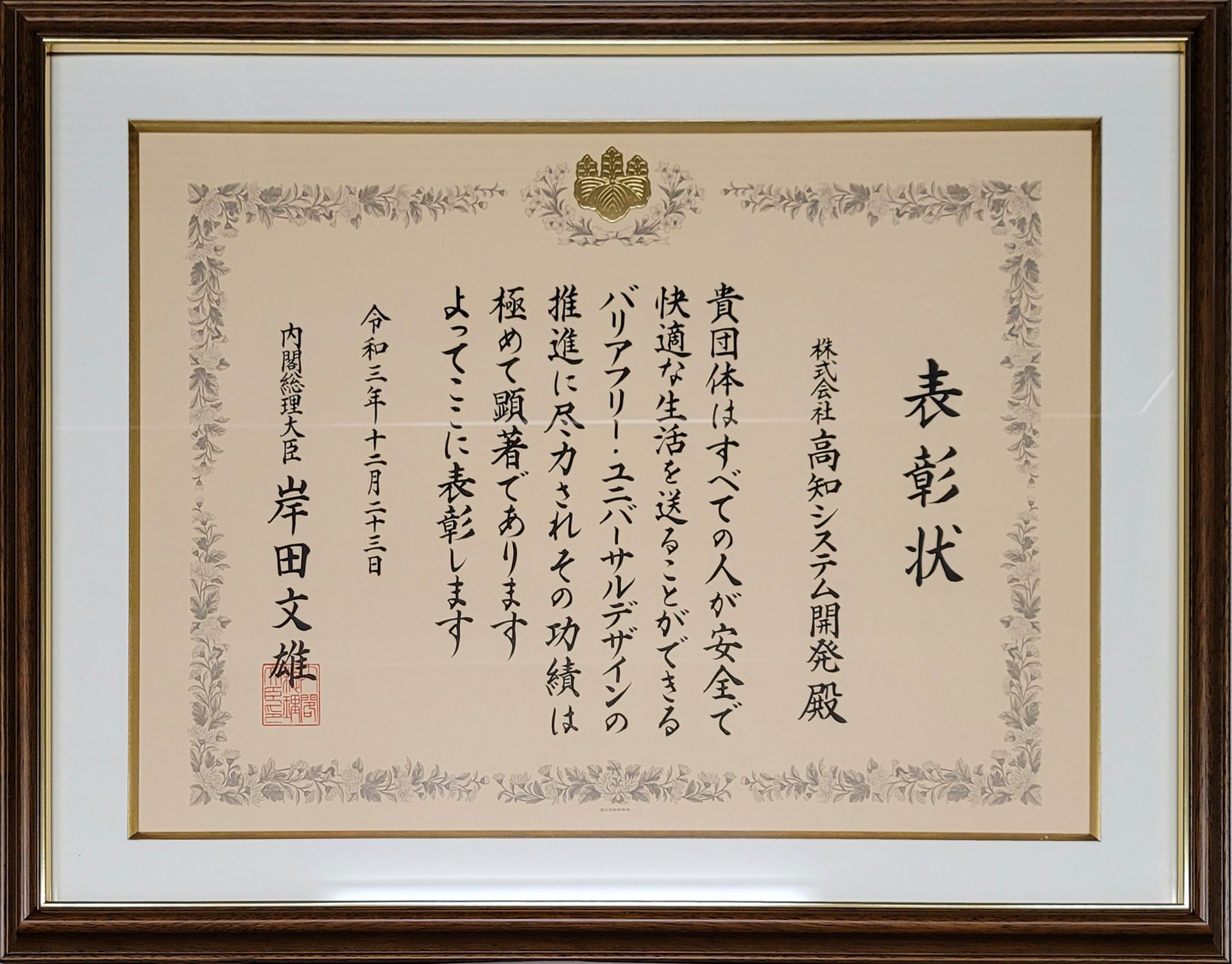 内閣総理大臣表彰の表彰状の写真。木製の額に入った表彰状には、株式会社高知システム開発殿、貴団体はすべての人が安全で快適な生活を送ることができるバリアフリー・ユニバーサルデザインの推進に尽力されその功績は極めて顕著であります、よってここに表彰します、令和三年十二月二十三日 内閣総理大臣 岸田文雄 と記されている